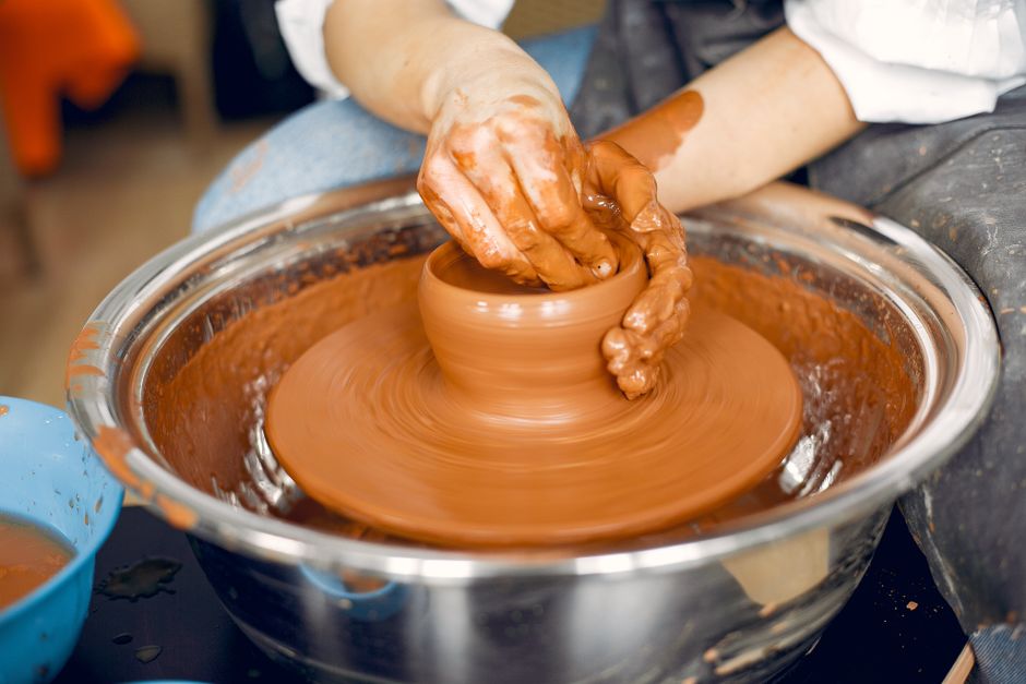 Crop anonymous craftswoman in apron and casual clothes making clay product on pottery wheel  in studio in daylight